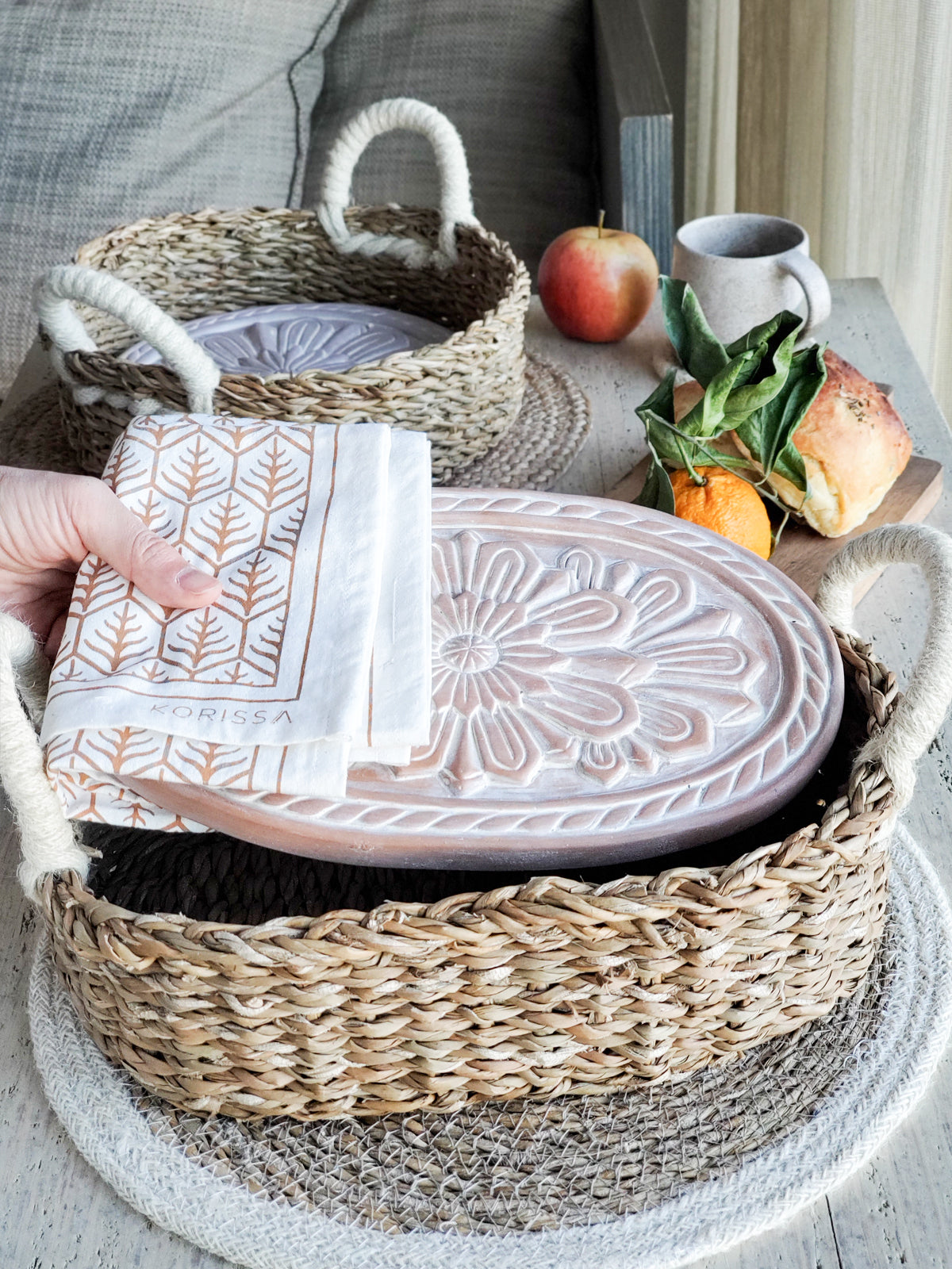 A decorative ceramic plate is beautifully set in a handwoven seagrass basket featuring handles, called the Bread Warmer & Basket Gift Set with Tea Towel - Flower. Below the basket are two patterned towels, crafted by Fair Trade artisans, showcasing leaf designs in black and orange.