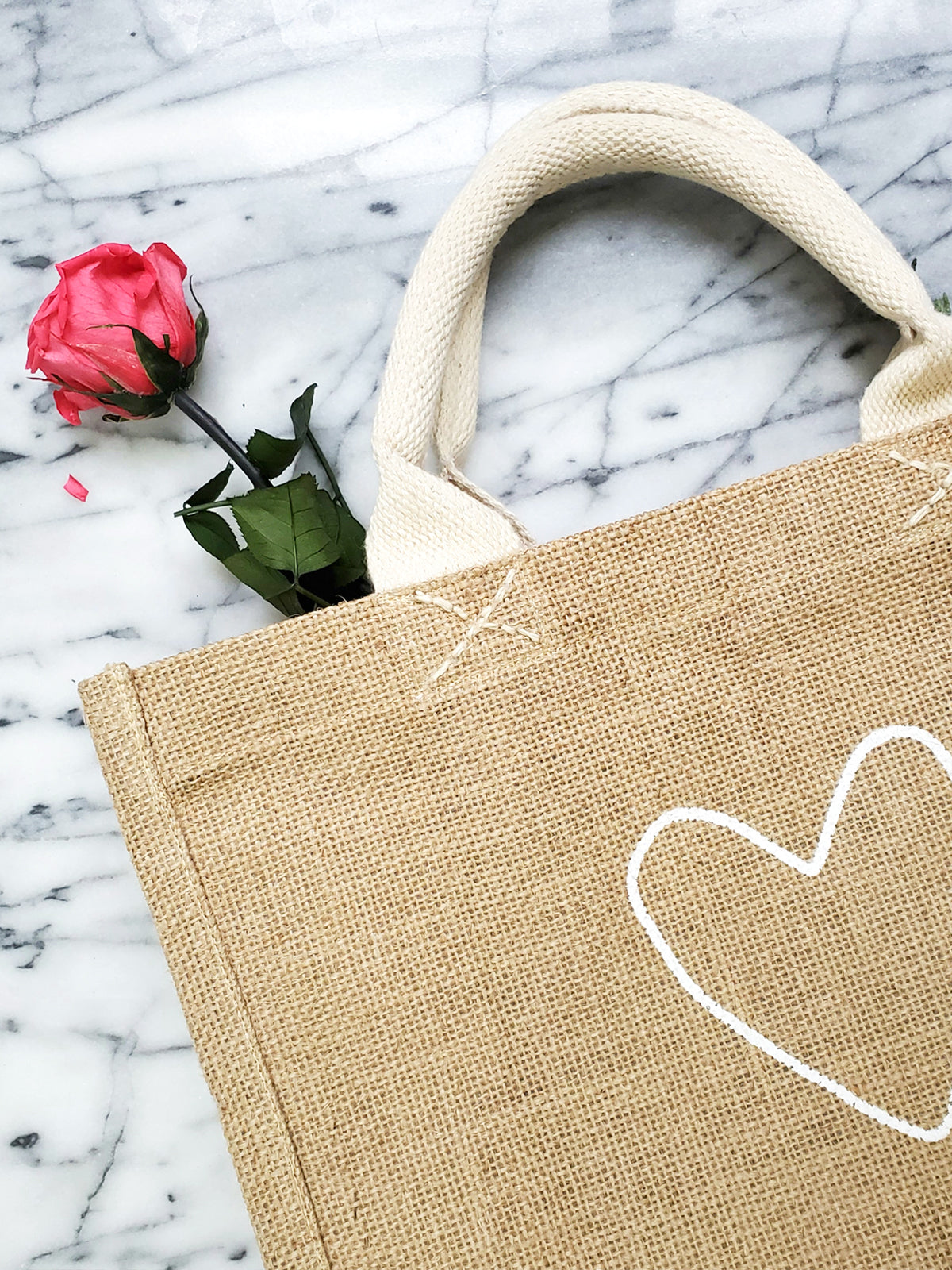 The "Gift Bag - Love" is a beige jute tote bag, handcrafted sustainably with short handles and adorned with a white heart outline on the front.