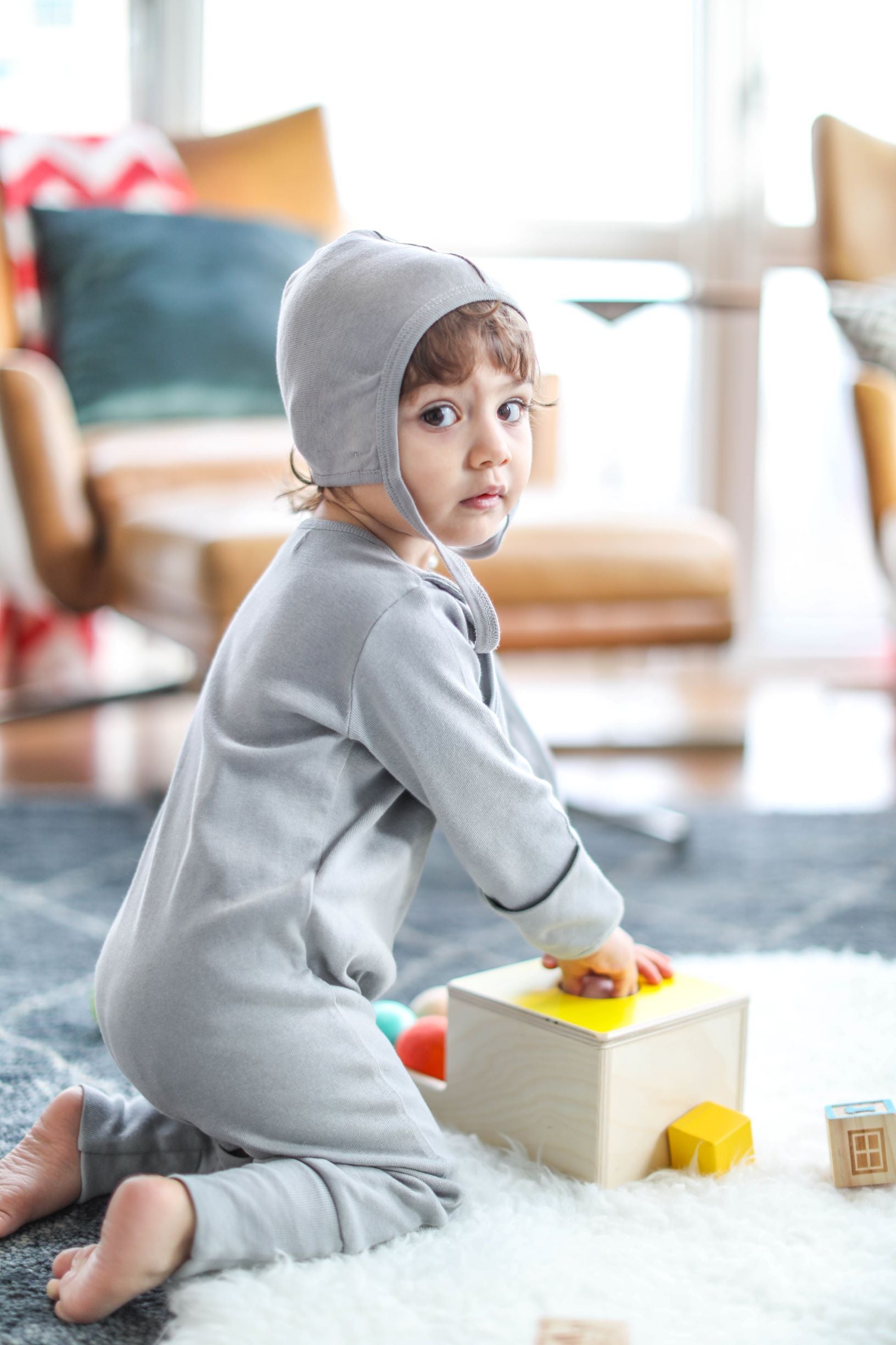 A baby is playing with the Smart Footed One-Piece + Bib - Gray by Fuchsia Milo in a living room.