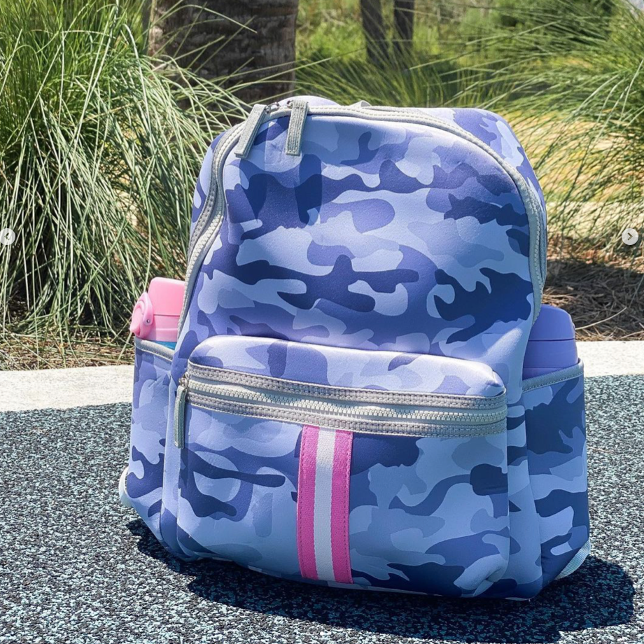 A woman in sunglasses, carrying a Neoprene Backpack designed for functionality, stands smiling on a gravel path amidst lush greenery, enjoying a laid-back outdoor environment.