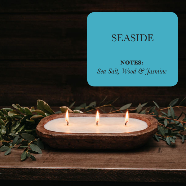 A luxurious 3-wick soy candle in a dough bowl sits on a table surrounded by lush greenery. Blue text above reads: "SEASIDE, Notes: Sea Salt, Wood & Jasmine.