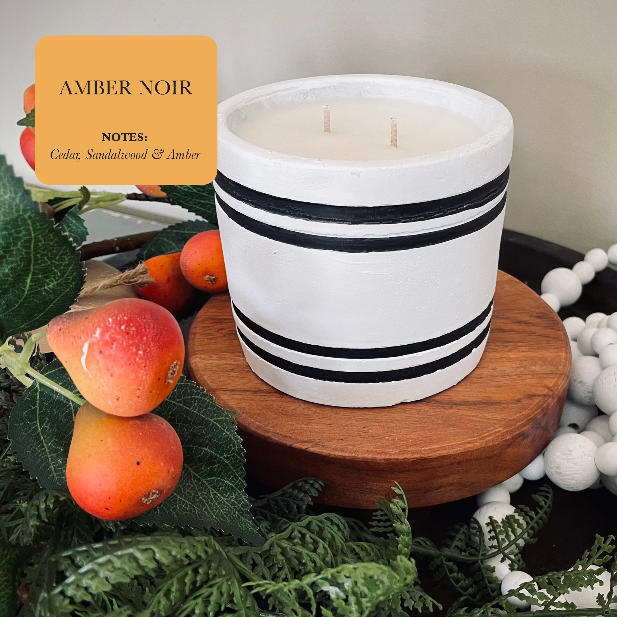 A luxury small striped stone designer candle labeled "Amber Noir" sits elegantly on a wooden stand, surrounded by artificial fruit and greenery. It adds a touch of sophistication to your home decor with its enticing notes of cedar, sandalwood, and amber.