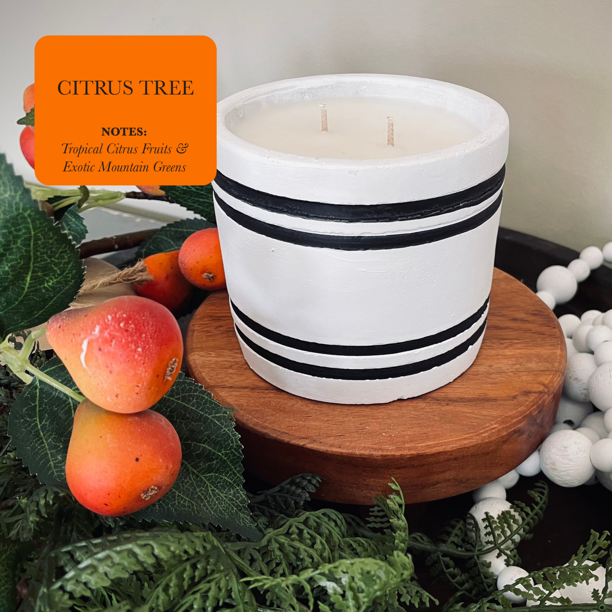 A Luxury Small Striped Stone Designer Candle - Citrus Tree, crafted with hand-poured natural soy wax, rests on a wooden surface adorned with green leaves and small orange fruits. An orange label displays "Citrus Tree," evoking the essence of tropical citrus fruits and exotic mountain greens.