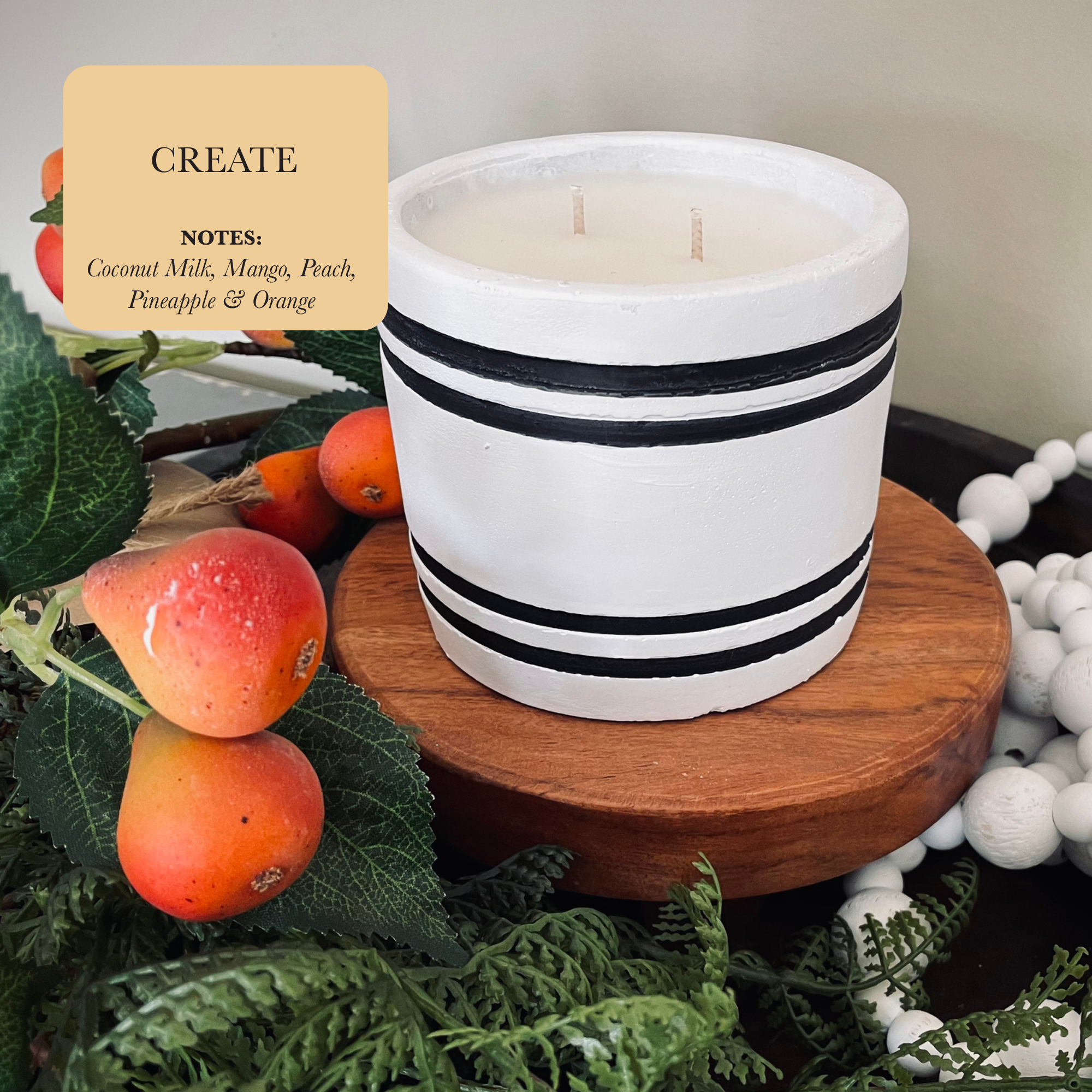 The Luxury Small Striped Stone Designer Candle - Create, with its black stripe design, sits gracefully on a wooden stand surrounded by green leaves and red berries. Its label reads: "CREATE NOTES: Coconut Milk, Mango, Peach, Pineapple & Orange," conjuring the essence of a tropical fruit paradise.