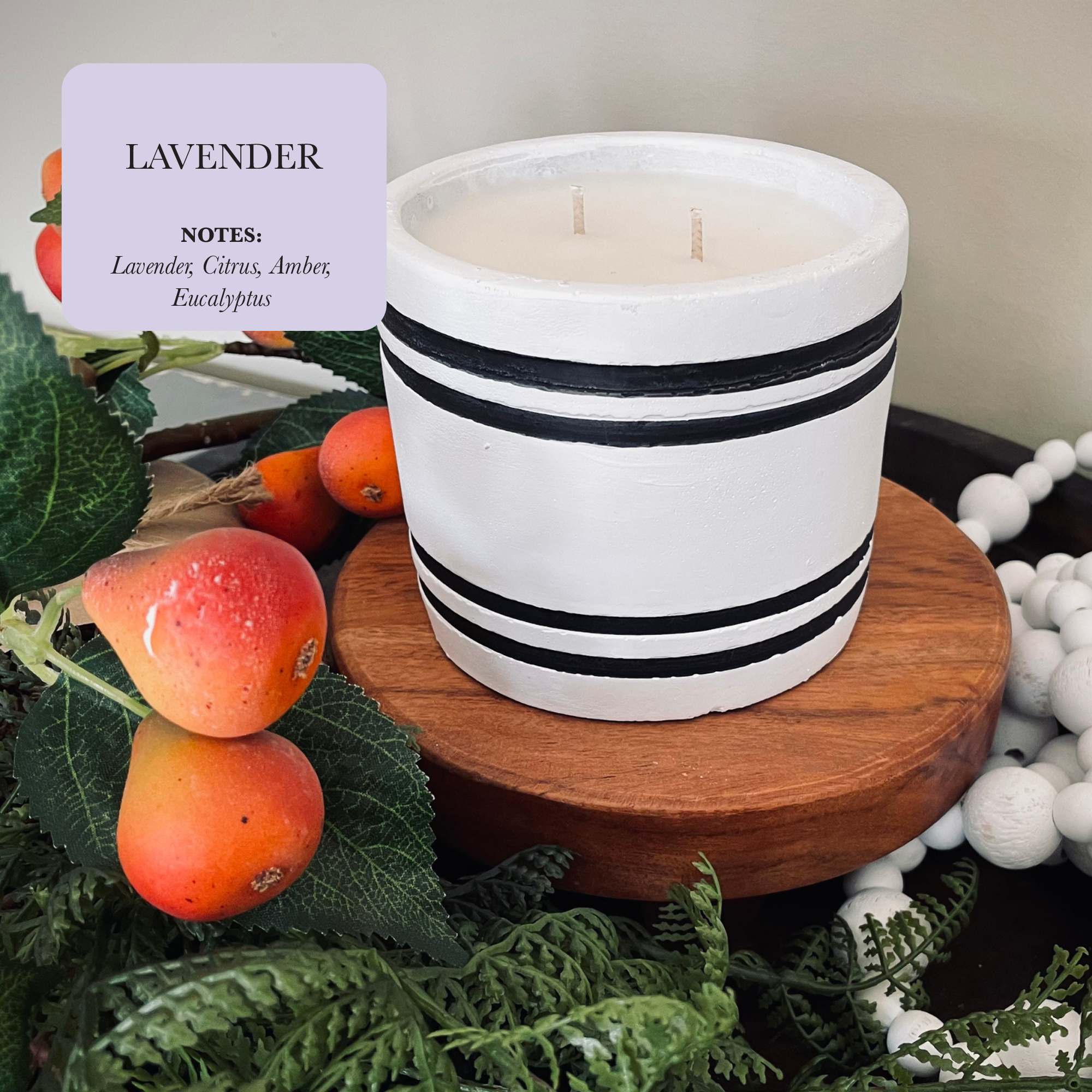 A Luxury Small Striped Stone Designer Candle in a white hue with black stripes rests on a wooden surface, enveloped by lush greenery and small round orange fruits. Text displays: "LAVENDER NOTES: Lavender, Citrus, Amber, Eucalyptus." Ideal for stress relief and relaxation.