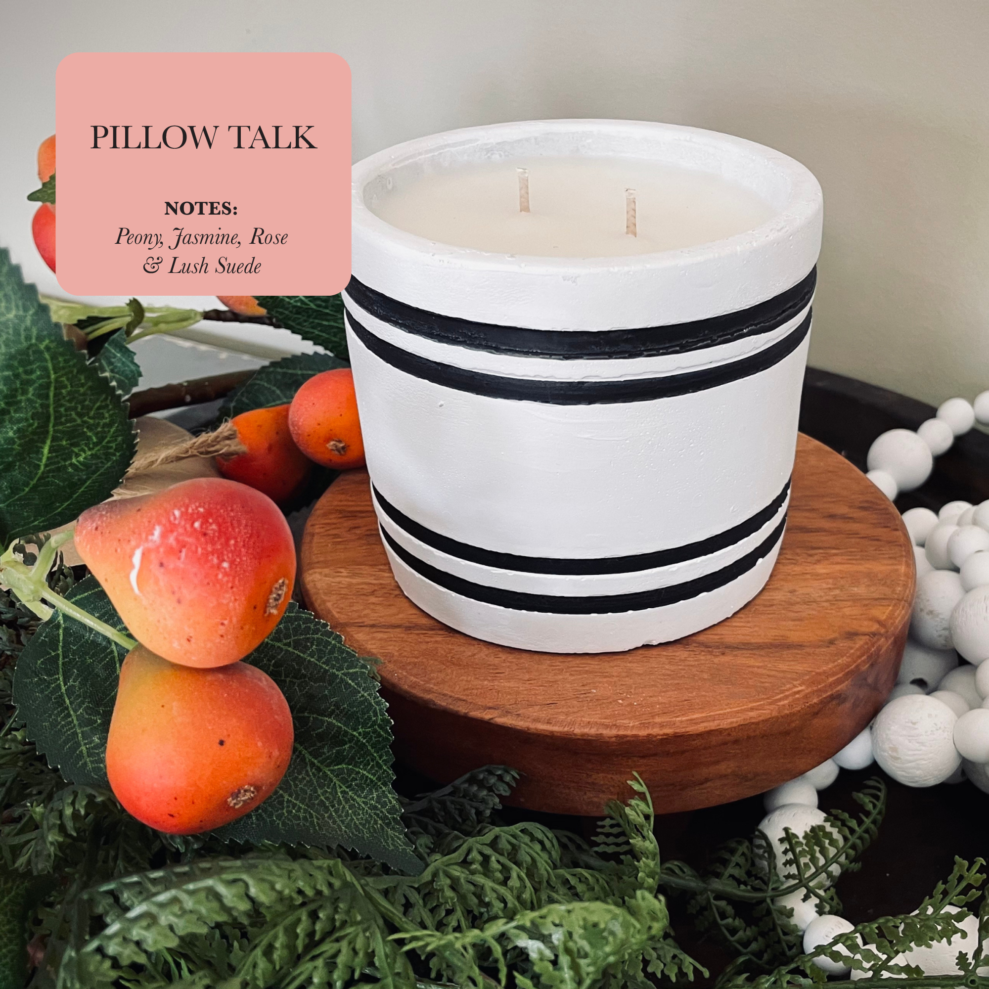 A Luxury Small Striped Stone Designer Candle - Pillow Talk with black stripes rests on a wooden surface surrounded by greenery and fruits. The label displays the name "Pillow Talk" and highlights the scent notes of jasmine, peony, rose, and lush suede.