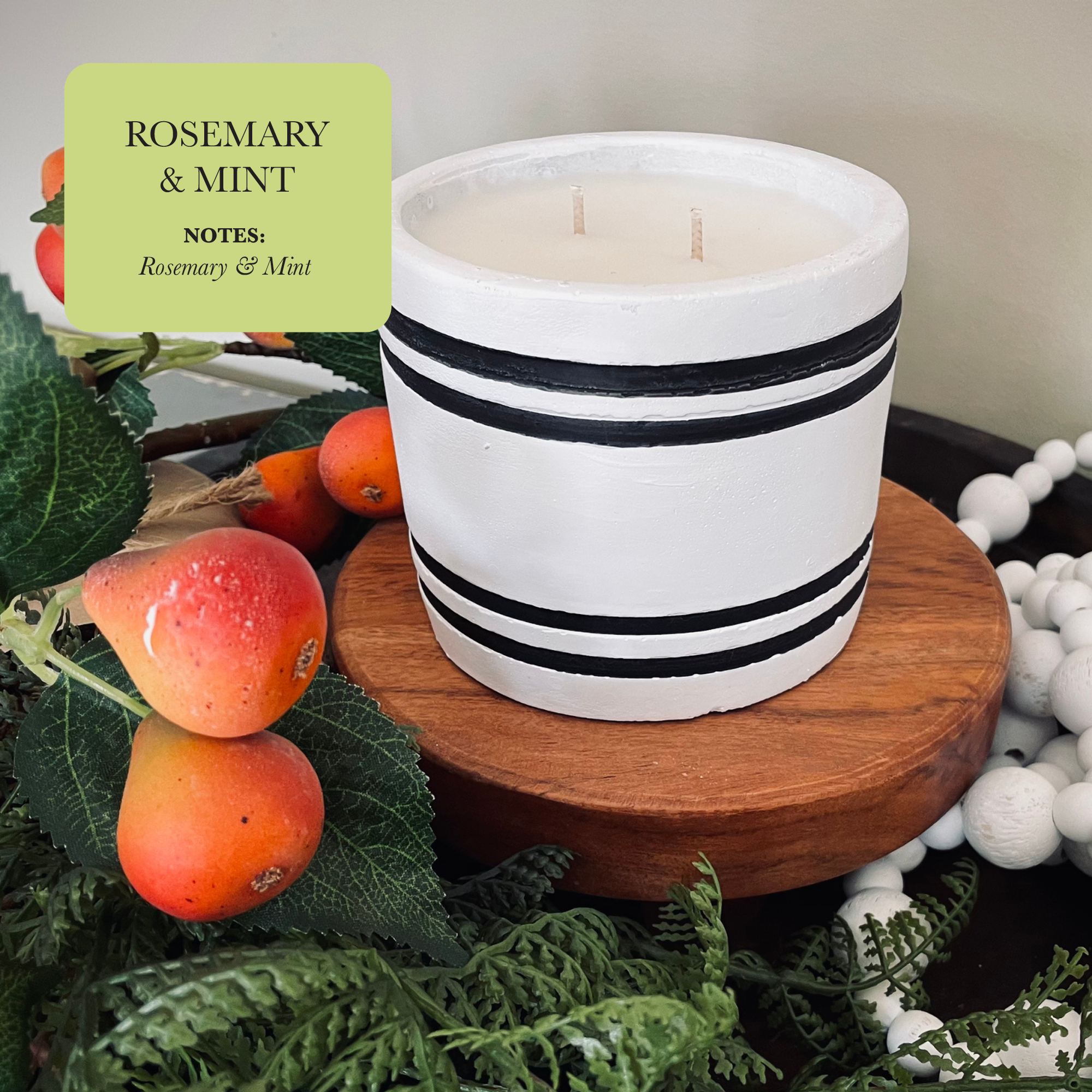 A hand-poured Luxury Small Striped Stone Designer Candle in Rosemary & Mint fragrance, featuring black stripes and placed on a wooden stand, is enhanced by surrounding greenery and vibrant red-orange fruits. Crafted from natural soy wax.