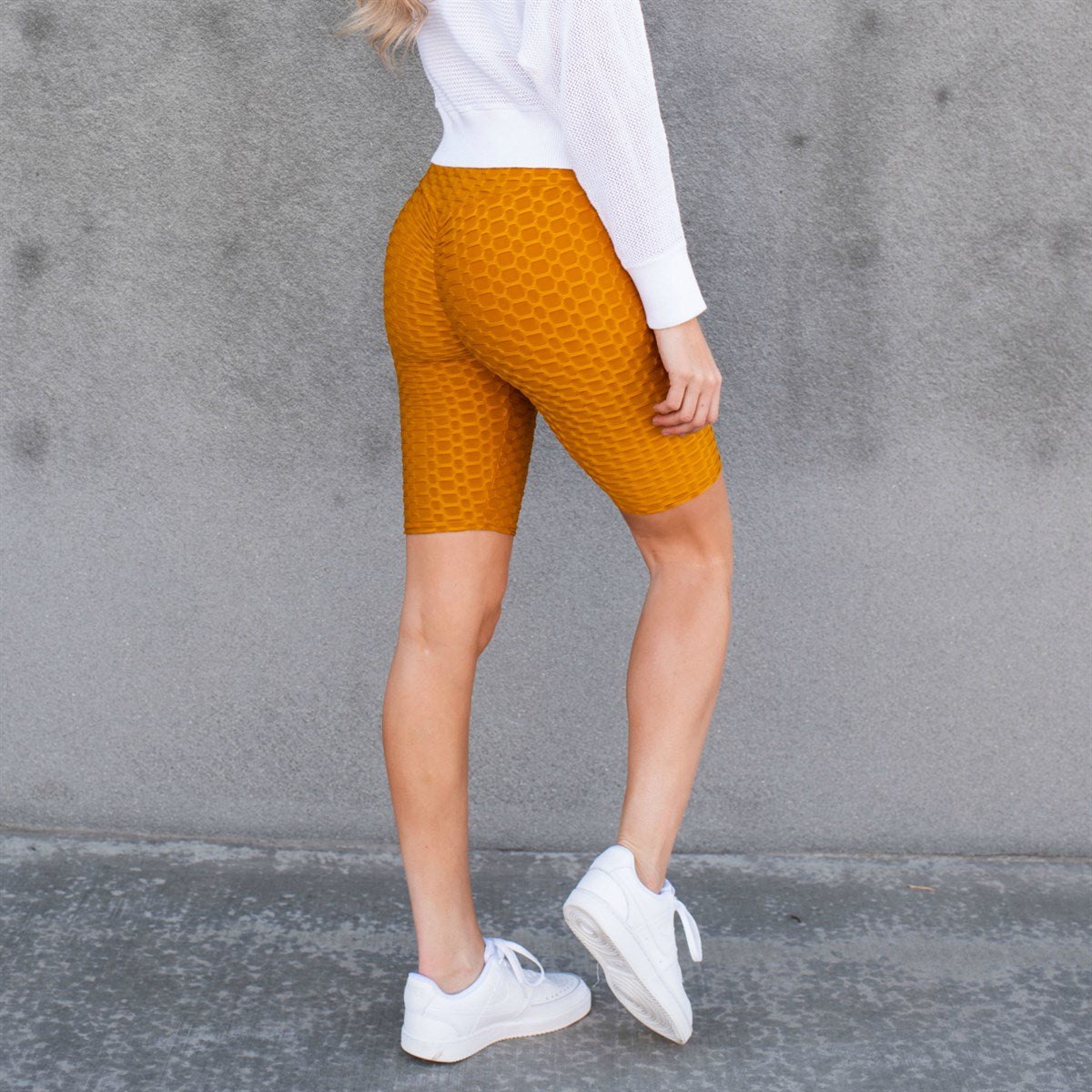 A person stands against a wall wearing a knotted white t-shirt, Cellulite Hiding Bermuda Shorts by New Mix showcasing a slim silhouette, and white sneakers. The face is out of the frame.