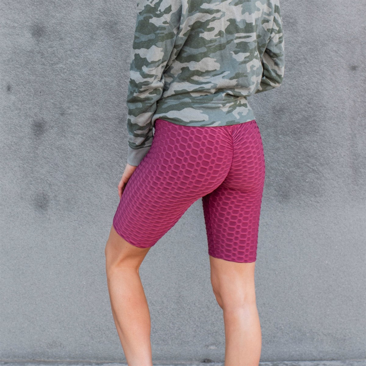 A person stands against a wall wearing a knotted white t-shirt, Cellulite Hiding Bermuda Shorts by New Mix showcasing a slim silhouette, and white sneakers. The face is out of the frame.