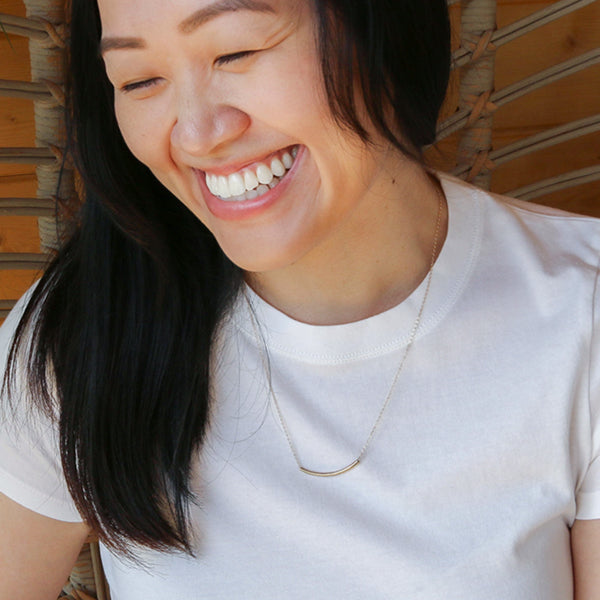 Amidst lush greenery, a person smiles outdoors, wearing a floral shirt and the delicate Talitha Necklace in 14k gold fill.