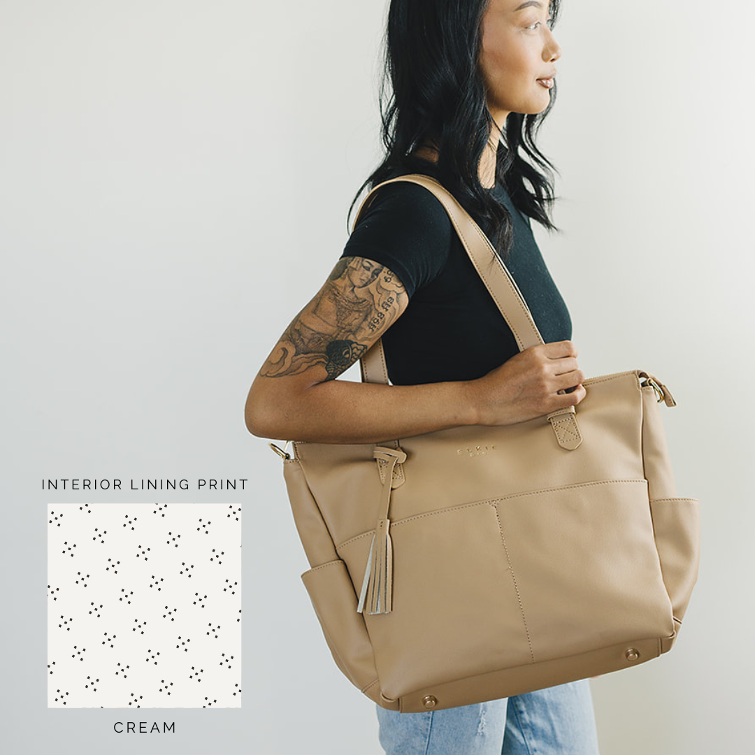 A person stands against a white brick wall, showcasing *The Aberdeen*, a stylish brown vegan leather tote with two front pockets and tassels.