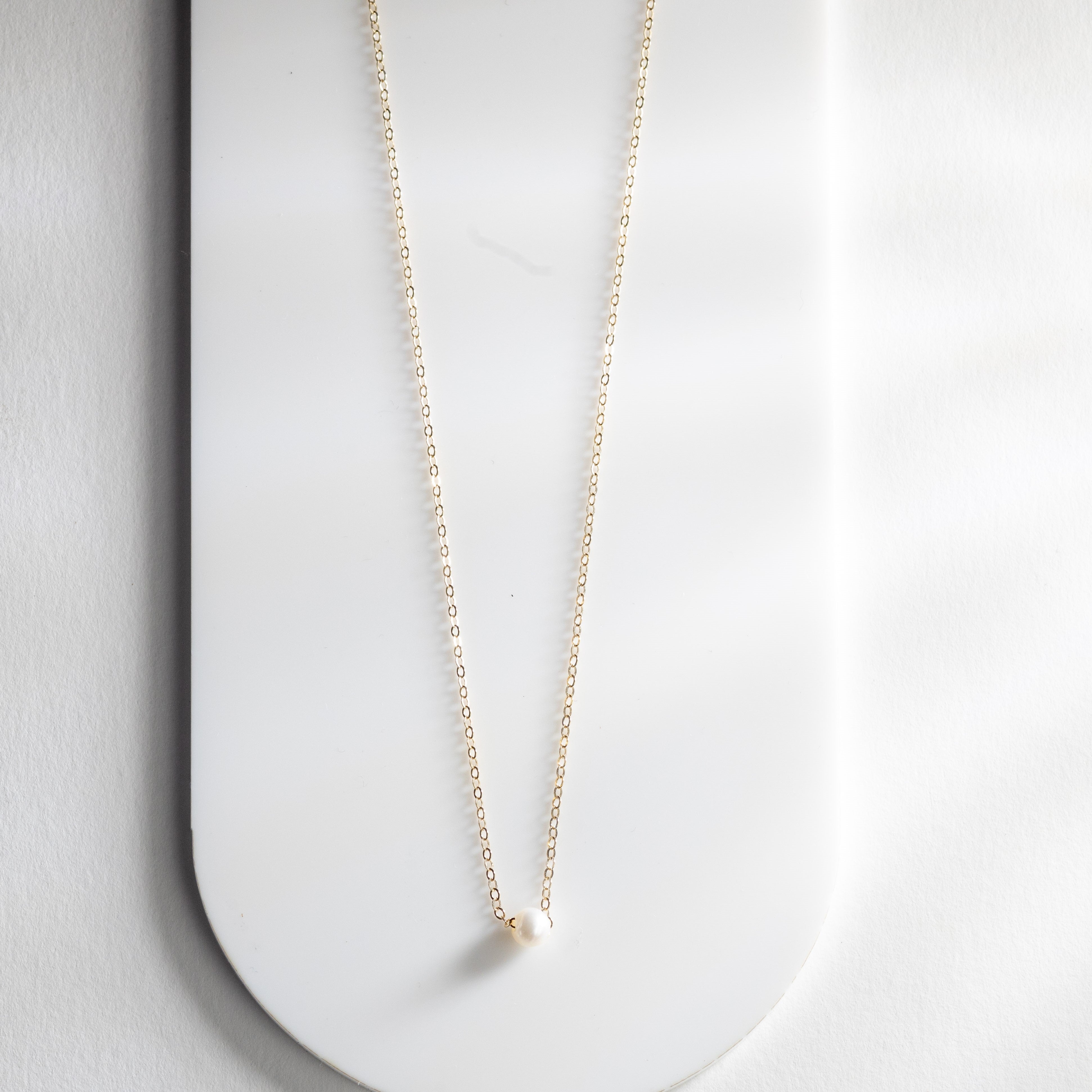 The Malaya Necklace, featuring a single small Philippine freshwater pearl pendant, is elegantly displayed on a white surface with its delicate 14k-gold filled chain.