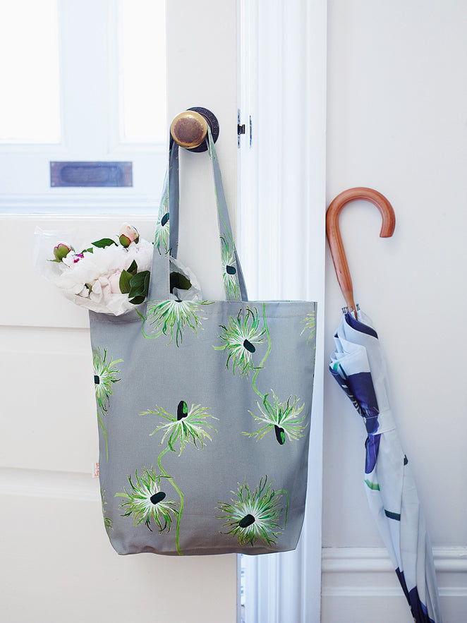 The "Thistle on Grey" tote bag, featuring a gray canvas adorned with floral patterns, hangs elegantly on a doorknob. This stylish shopping tote is filled with pink and white flowers, evoking the charm of an oil-painted scene. Nearby, an umbrella leans against the wall, adding to the picturesque arrangement.