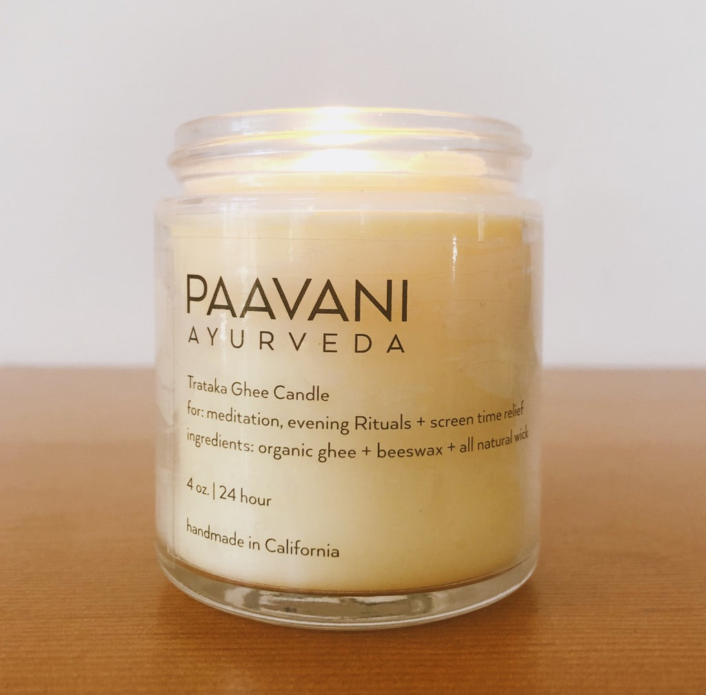 The Trataka Ghee Candle, encased in a glass jar labeled "PAAVANI," features a yellow balm crafted with turmeric, coconut wax, and essential oils. Inspired by Trataka practices, its soothing ingredients are designed to potentially enhance eye health.