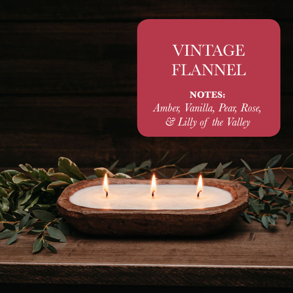 A luxury 3-Wick Dough Bowl Soy Candle - Vintage Flannel, featuring notes of amber, vanilla, pear, rose, and lily of the valley, is nestled amidst lush green foliage. This flannel-scented candle emits a warm and inviting aroma, wrapping you in comfort with its wax embrace.