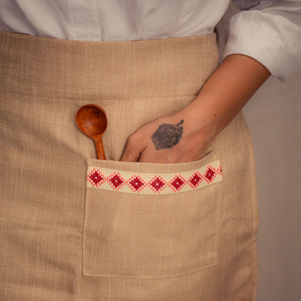 A person wearing a cream Farah Half Apron with a wooden spoon tucked into the front pocket. This apron showcases an empowering red and white embroidered tatreez motif.