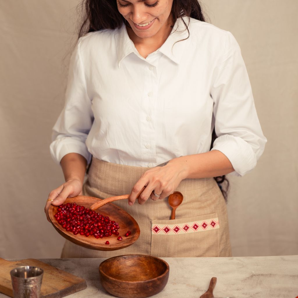 A person wearing a cream Farah Half Apron with a wooden spoon tucked into the front pocket. This apron showcases an empowering red and white embroidered tatreez motif.