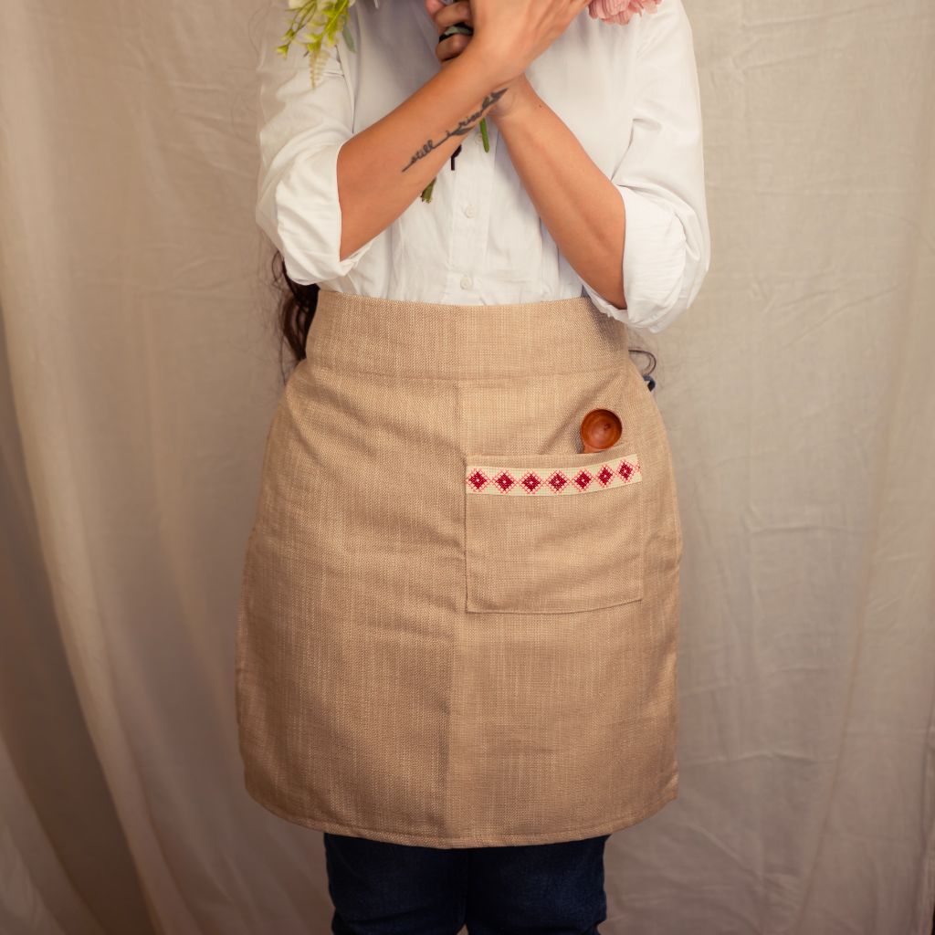 A person wearing a cream Farah Half Apron with a wooden spoon tucked into the front pocket. This apron showcases an empowering red and white embroidered tatreez motif.