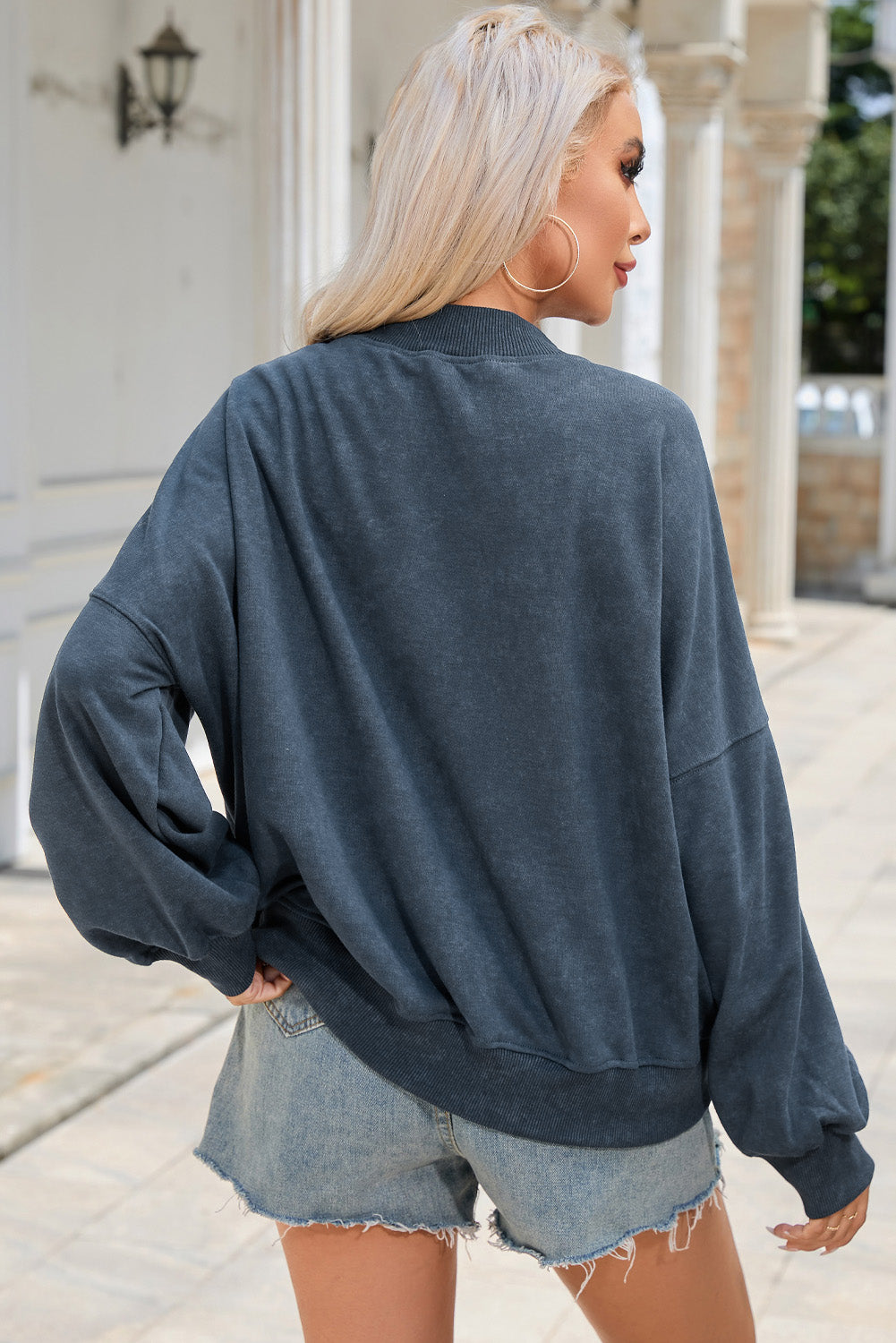 A person with long blonde hair is wearing a sky blue drop shoulder crew neck pullover sweatshirt and light denim shorts, standing outdoors with their back to the camera.