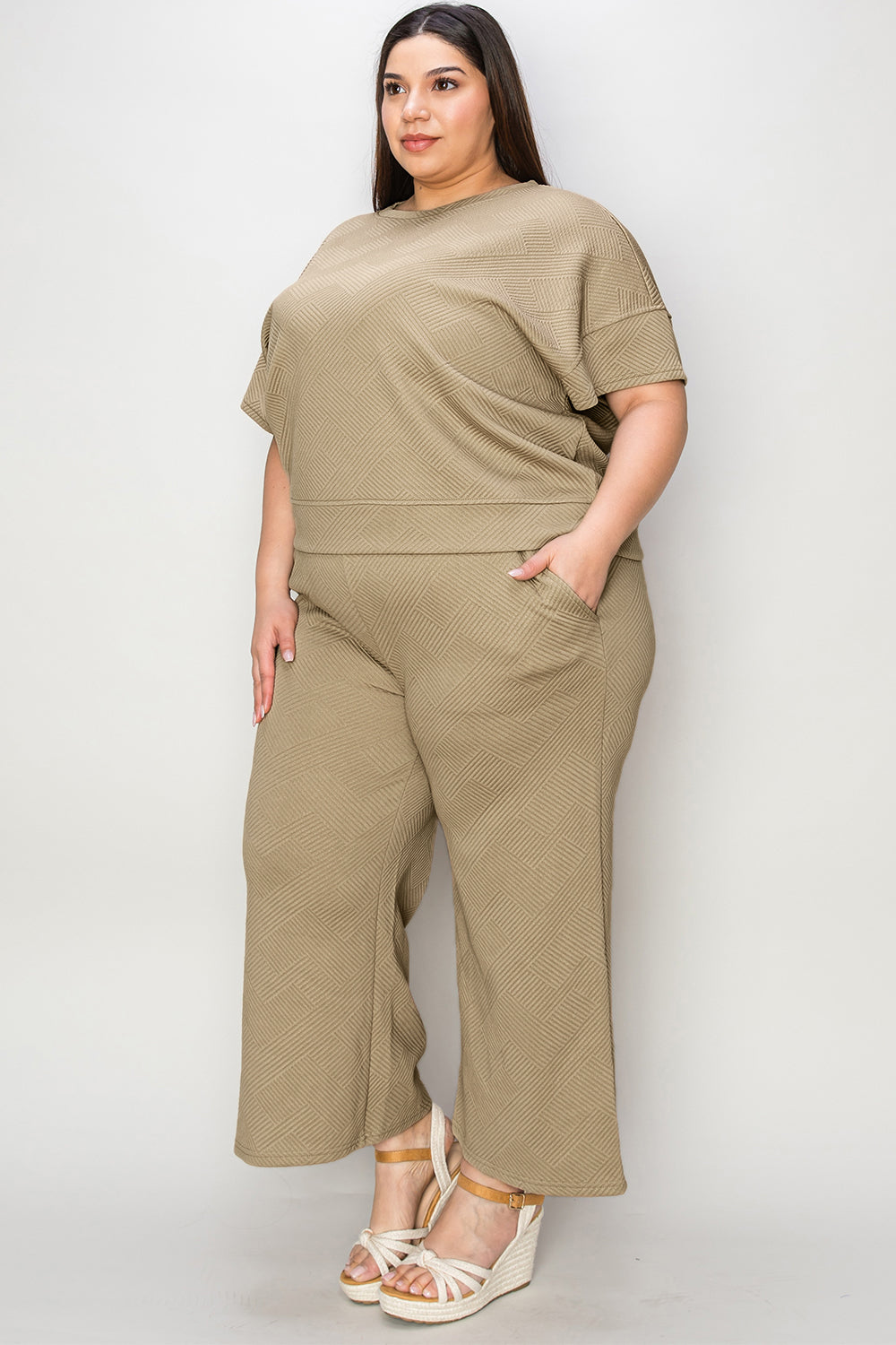 A woman with long hair smiles while standing outside a building, dressed in the Double Take Full Size Texture Short Sleeve Top and Pants Set, featuring a matching beige two-piece ensemble. She wears slides and holds a hat in her right hand. Her outfit, slightly stretchy for added comfort, is perfect for machine wash cold.