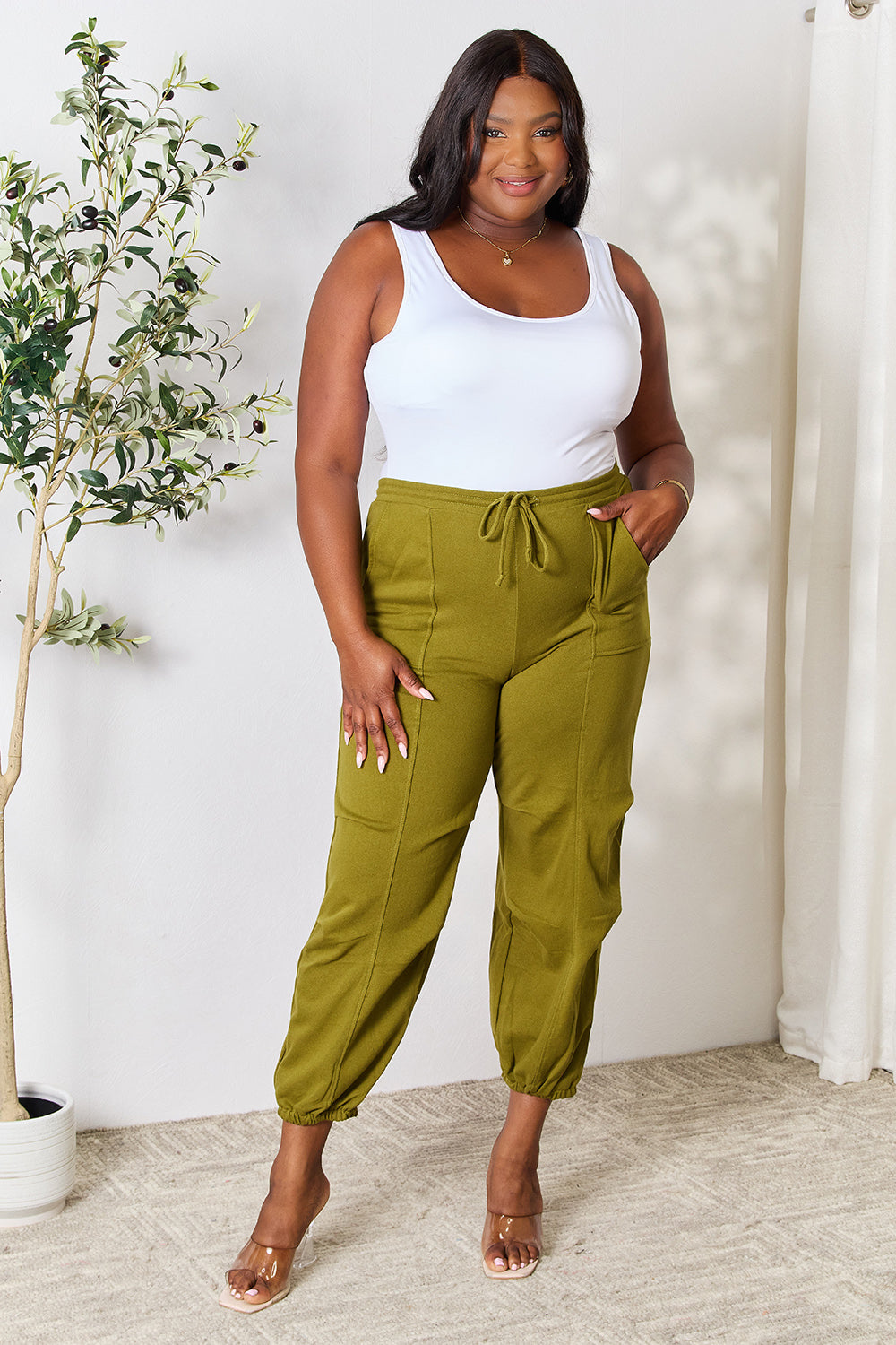 A person wearing Culture Code Full Size Drawstring Sweatpants with pockets in olive green and a white sleeveless top stands on a light-colored carpet.