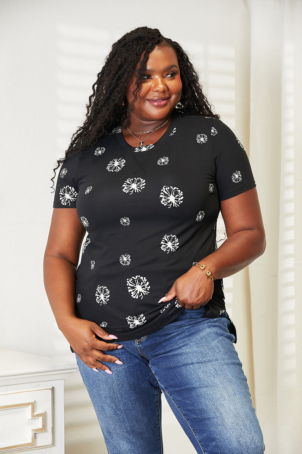 A woman with long braided hair is smiling and wearing the Double Take Dandelion Print Round Neck T-Shirt paired with blue jeans. She stands in front of a light-colored wall, embodying nature-inspired fashion.