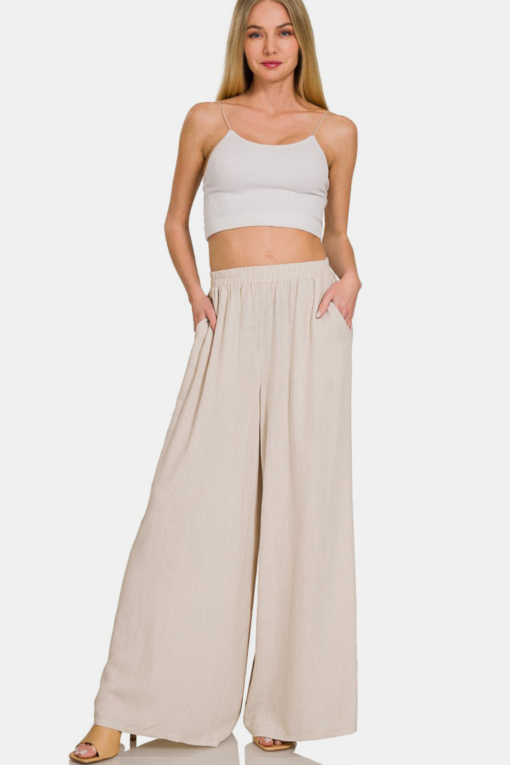 Person wearing Zenana Pleated Linen Blend Wide Leg Pants in beige with hands in pockets, shown from waist to feet. The pleated pants have an elastic waistband and flowy fabric. Model is also wearing light-colored open-toed sandals.