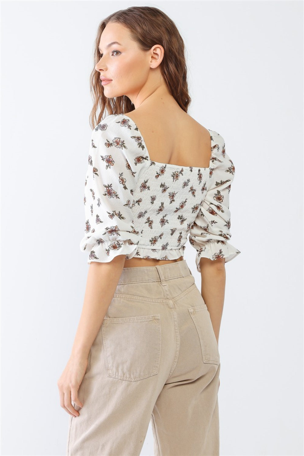 A woman with long hair poses in the PAPERMOON Floral Ruffled Smocked Crop Top, elegantly paired with light beige pants against a plain background.