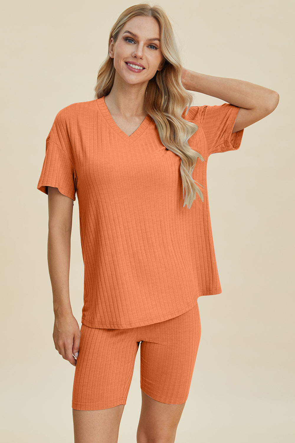 A woman with long blonde hair wearing the Basic Bae Full Size Ribbed V-Neck Short Sleeve Top and Shorts Set in rust stands against a plain background. This stylish two-piece set is made from a comfortable, stretchy fabric that is also machine washable.