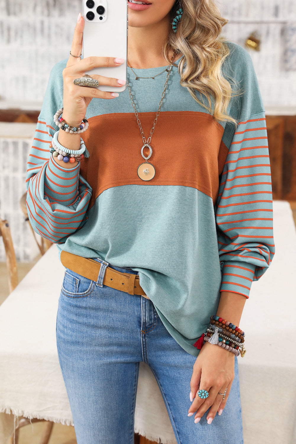 A woman wearing a Green Colorblock Striped Bishop Sleeve Top with trendy side slits and a flattering fit, paired with blue jeans, accessorized with layered necklaces, bracelets, and rings, is taking a selfie with a smartphone.