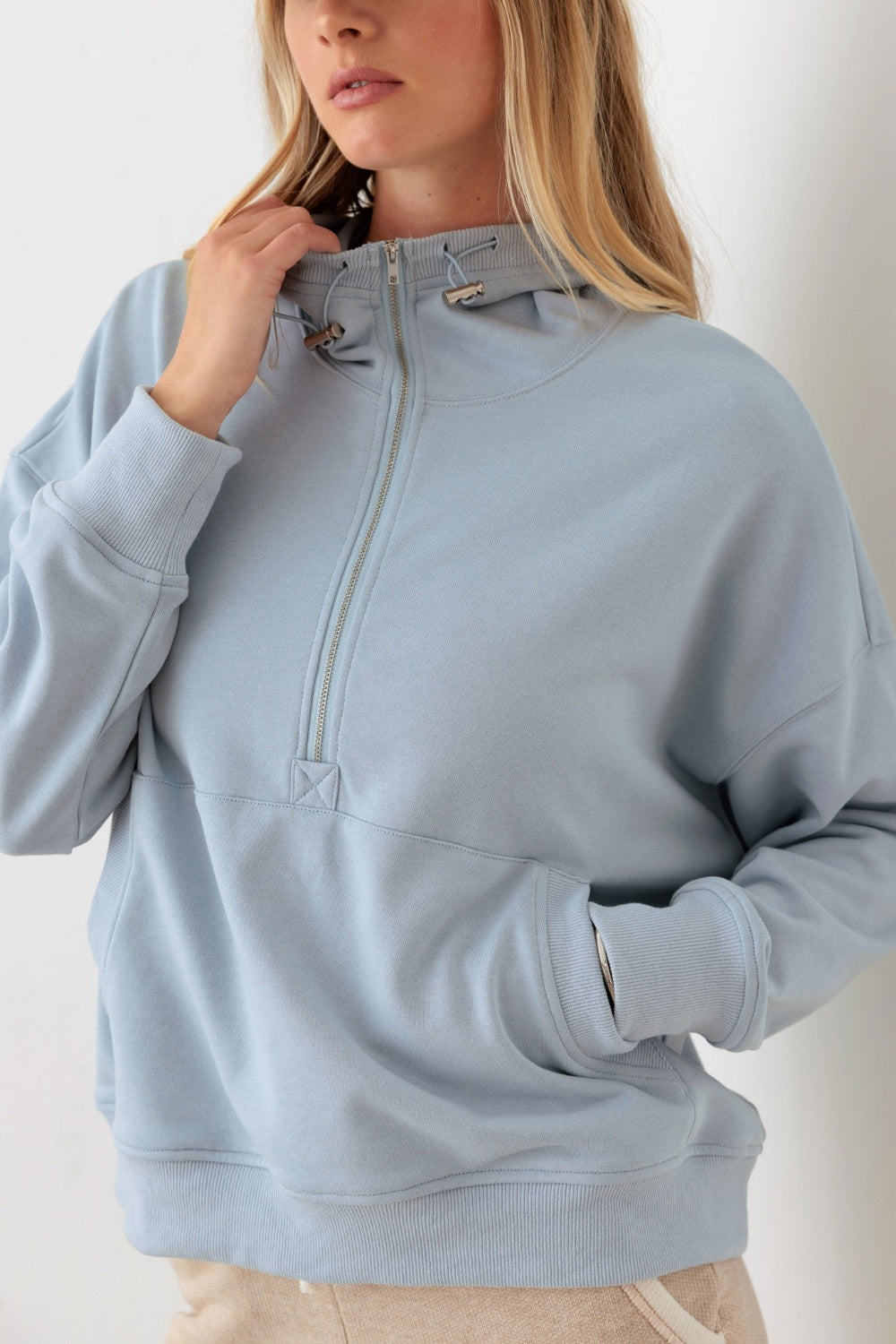 In a neutral-colored room, a person is wearing the Le Lis Half Zip Drawstring Mock Neck Hoodie in light blue along with a beige cap.