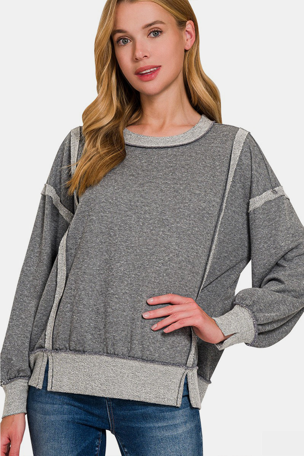 A woman with long blonde hair is seen in a neutral pose against a plain white background, wearing the grey Zenana Washed Exposed-Seam Sweatshirt featuring a crew neck and ribbed hem. Her casual wear is perfect for layering in any season.