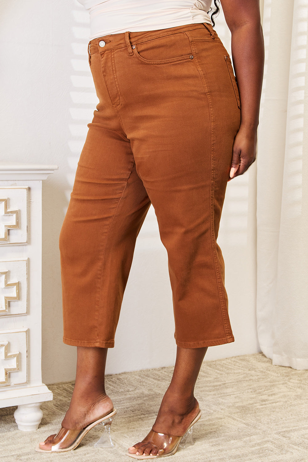 A chic and modern individual stands near a white dresser in a sunlit room, wearing stylish Judy Blue Full Size Tummy Control Garment Dyed Wide Crops and clear-heeled sandals.