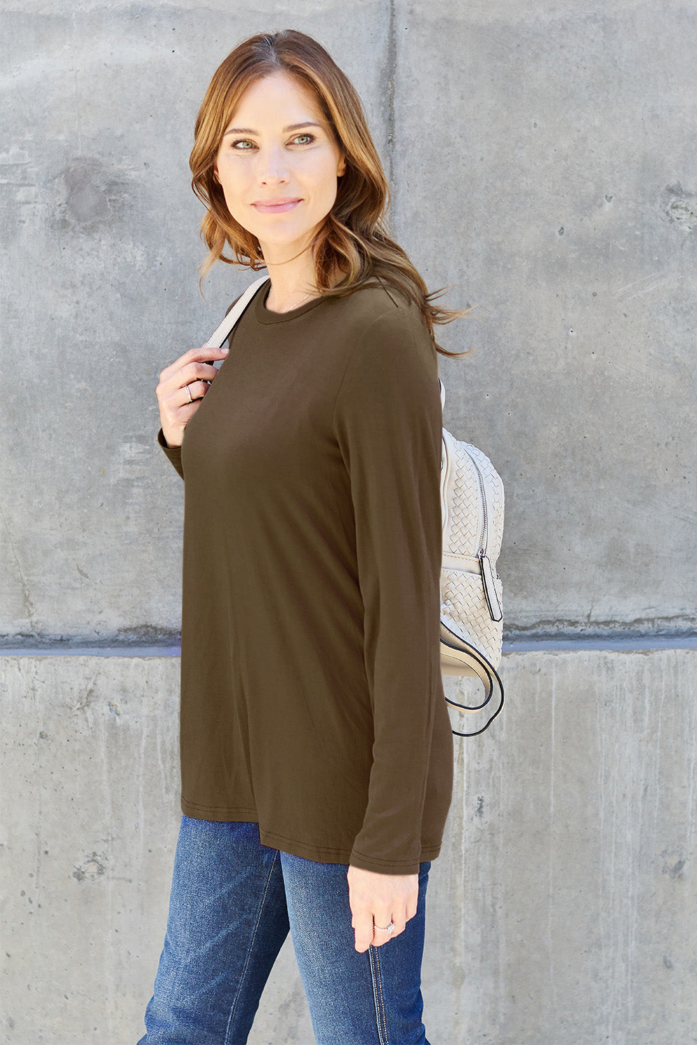 A woman with shoulder-length hair wearing the Basic Bae Full Size Round Neck Long Sleeve Top and blue jeans stands in front of a concrete wall, holding a beige handbag over her shoulder. Her outfit exemplifies basic style, with the top made from 100% polyester and recommended for machine wash cold.