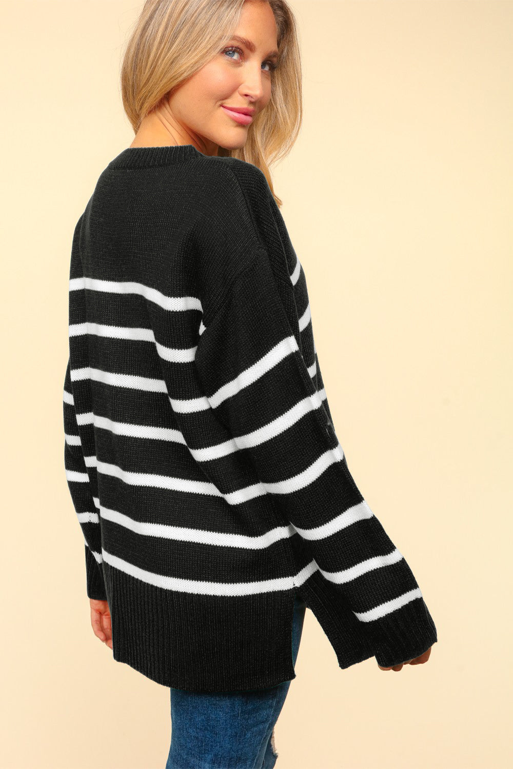 A person with long blonde hair is wearing the Haptics Full Size Striped Contrast Side Slit Sweater paired with blue jeans, standing against a plain beige background.