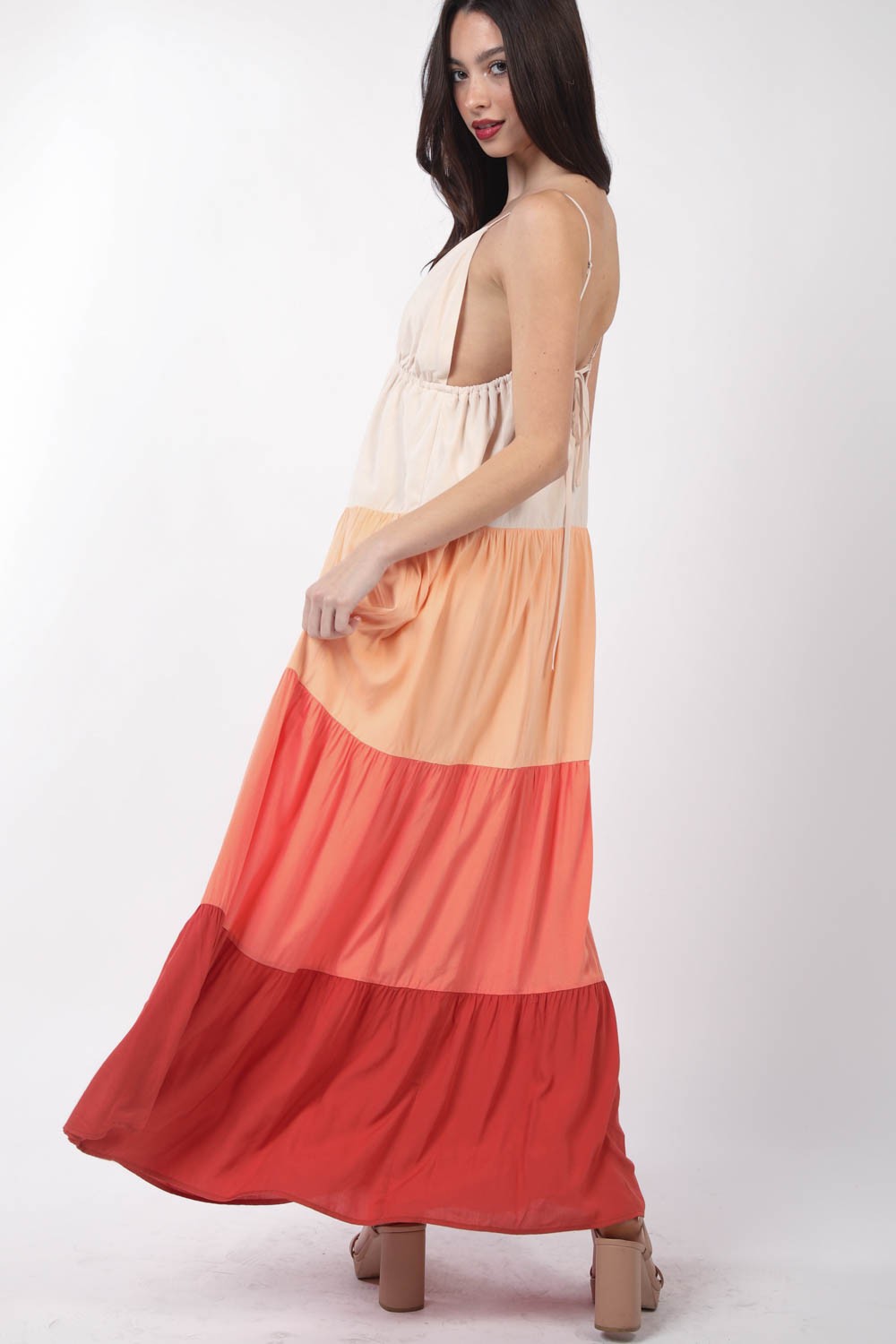 A woman stands, wearing the VERY J Color Block Tiered Maxi Cami Dress in shades of beige, peach, and red, paired with beige sandals. Her right hand holds the side of her dress as she poses against a plain white background—a truly fashionable choice.