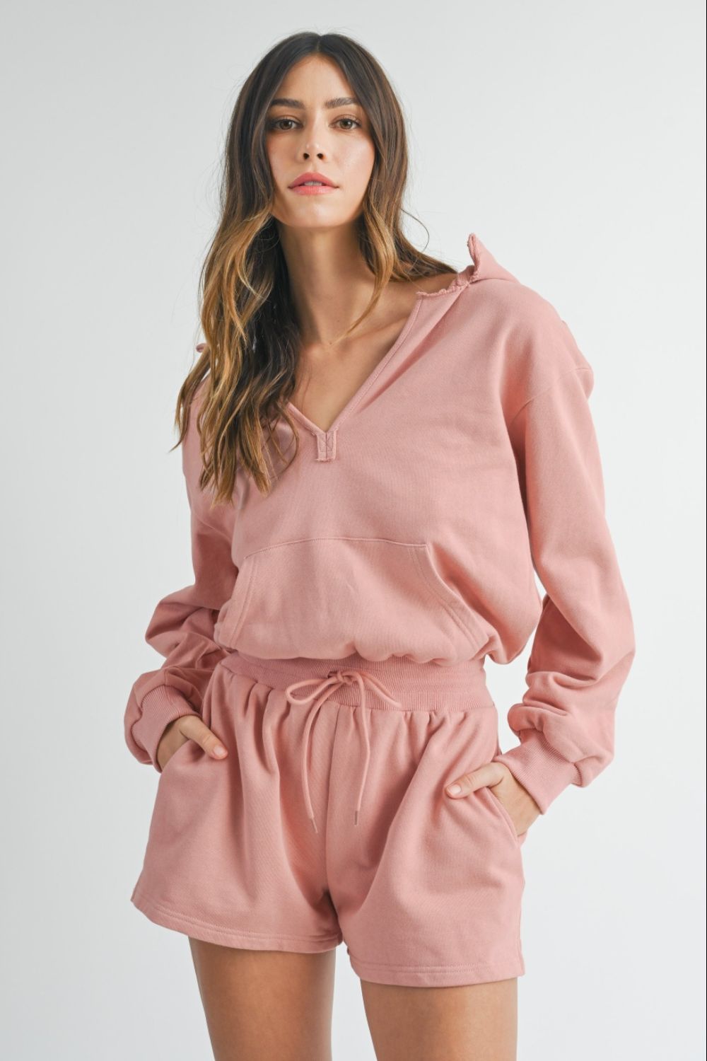 Woman wearing a MABLE French Terry Hooded Romper in pink, hands in pockets, looking directly at the camera.