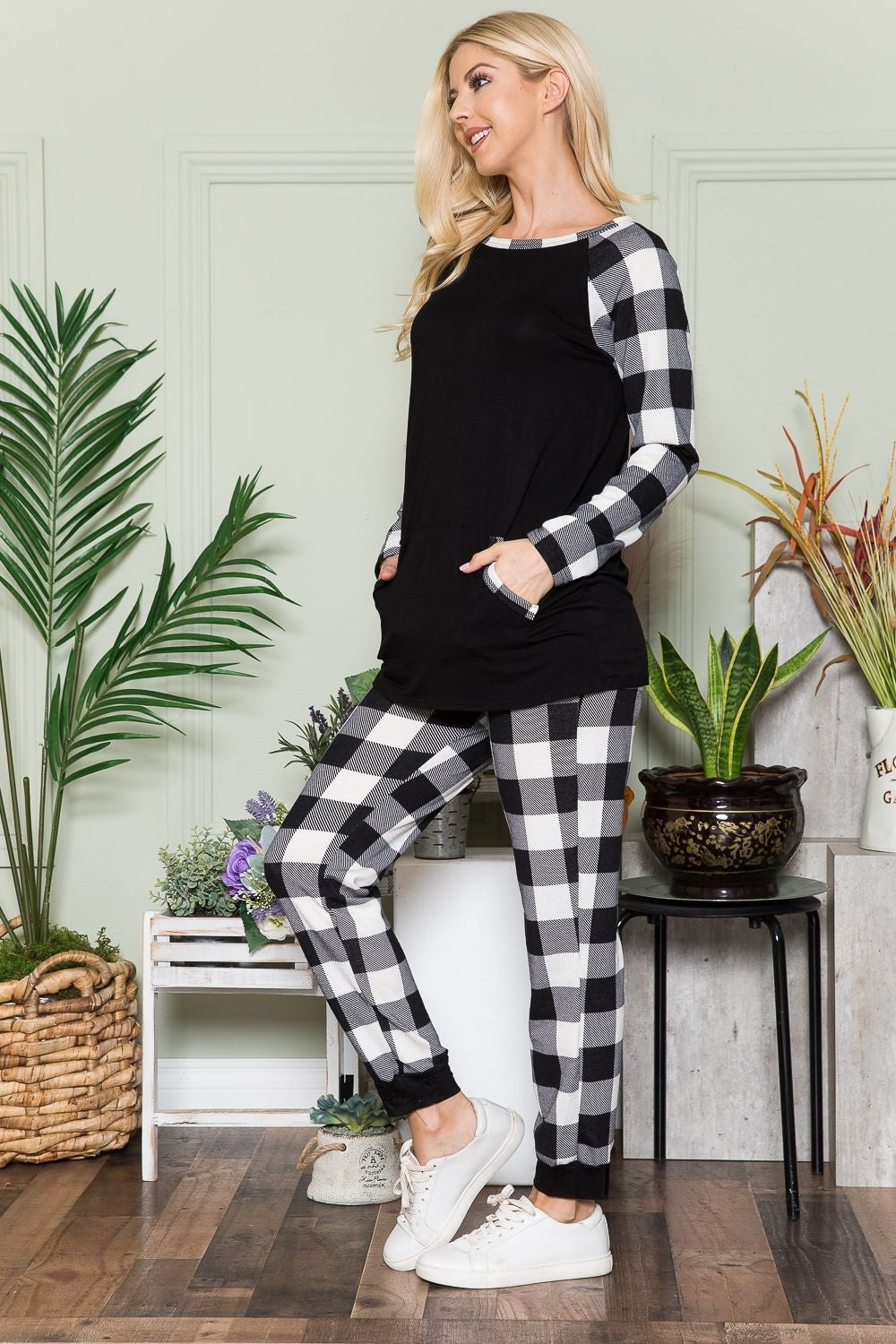 A person wearing the Celeste Plaid Long Sleeve T-Shirt with Pockets stands in a room filled with plants and wooden flooring, presenting a laid-back style.