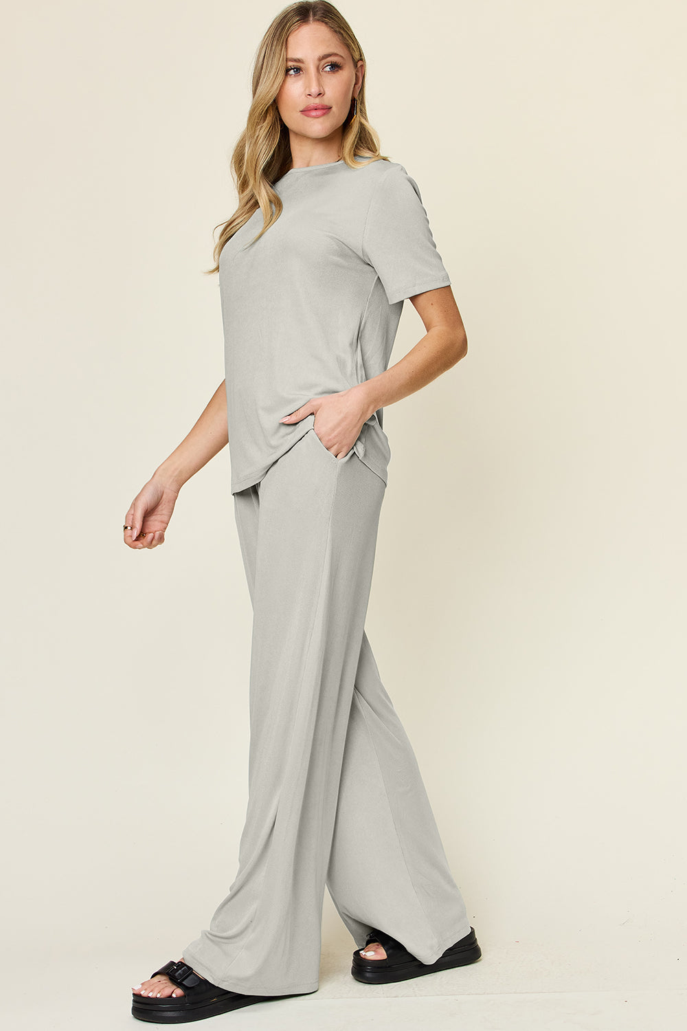 A person stands wearing the Double Take Full Size Round Neck Short Sleeve T-Shirt and Wide Leg Pants Set in a stretchy light grey fabric, with one hand in a pocket and black sandals.