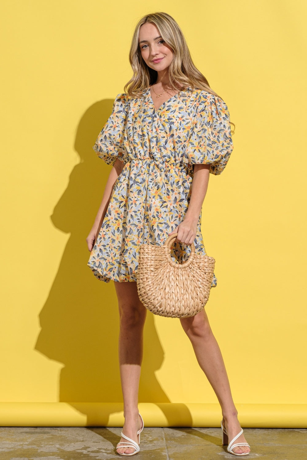 A person wearing the And The Why Full Size Floral Surplice Puff Sleeve Dress stands against a yellow background, holding a woven bag and wearing white sandals.