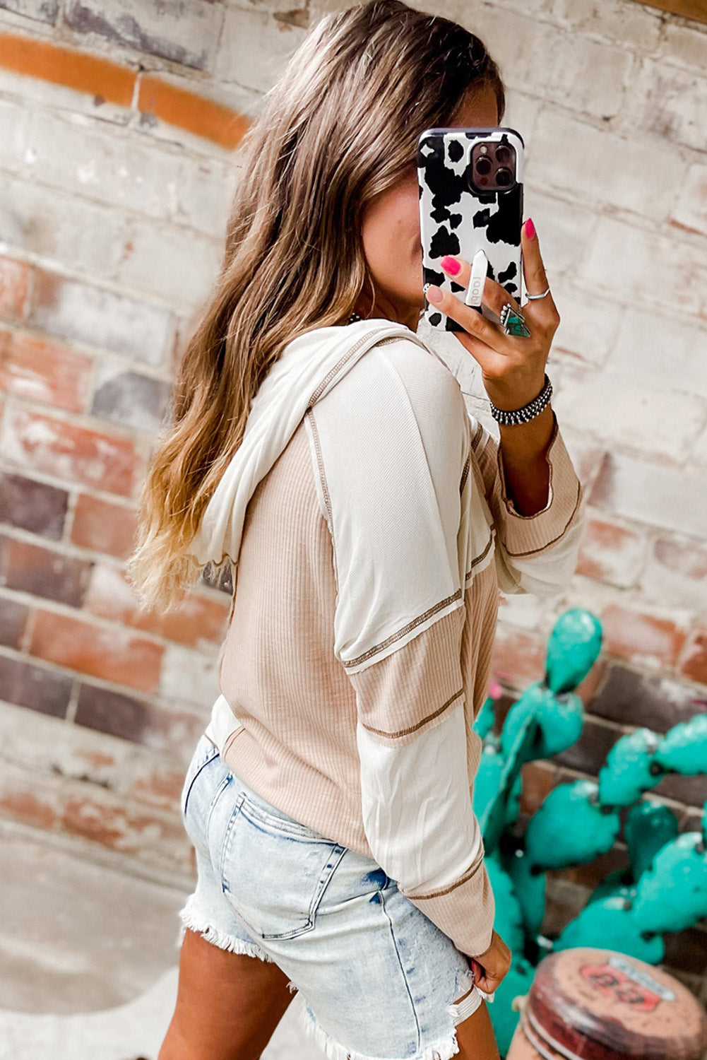 A person with light blonde hair in a bun, wearing sunglasses and a Multicolor Colorblock Seamed Ribbed Henley Hoodie, is walking outside on a paved street.