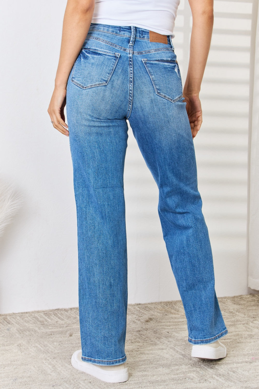 A person wearing the Judy Blue Full Size High Waist Distressed Straight-Leg Jeans and white sneakers stands against a white wall.