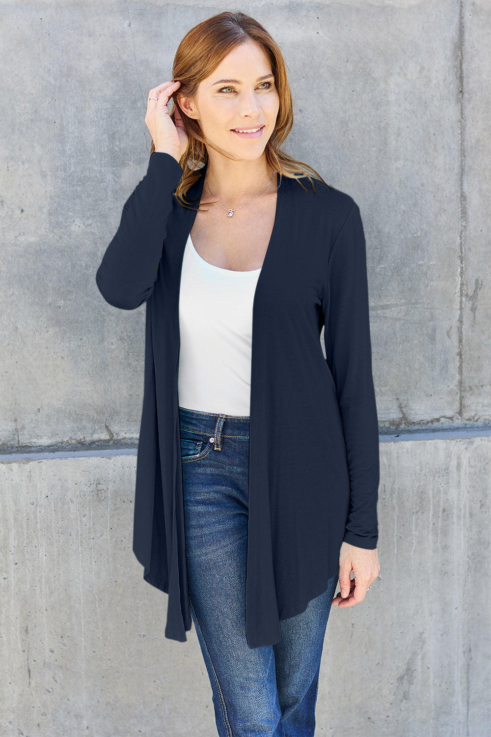 A woman wearing the Basic Bae Full Size Open Front Long Sleeve Cardigan in navy blue, paired with a white top and blue jeans, stands against a concrete wall. She touches her hair and gazes to the side, embodying a chic yet simple style. The slightly stretchy materials of the cardigan ensure comfort while maintaining its stylish simplicity. For easy care, machine wash it cold.