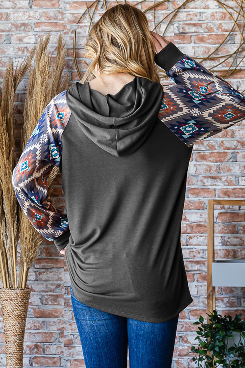 A woman wearing a Heimish Half Button Printed Long Sleeve Hooded Top in dark gray, showcasing patterned sleeves, stands in front of a brick wall, holding the hood with one hand and placing the other hand in her pocket.