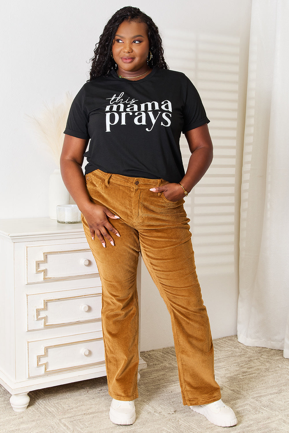 A woman wearing a Simply Love THIS MAMA PRAYS Graphic T-Shirt, paired with brown pants, stands gracefully in a softly lit room, exuding a sense of calm and spiritual gift.