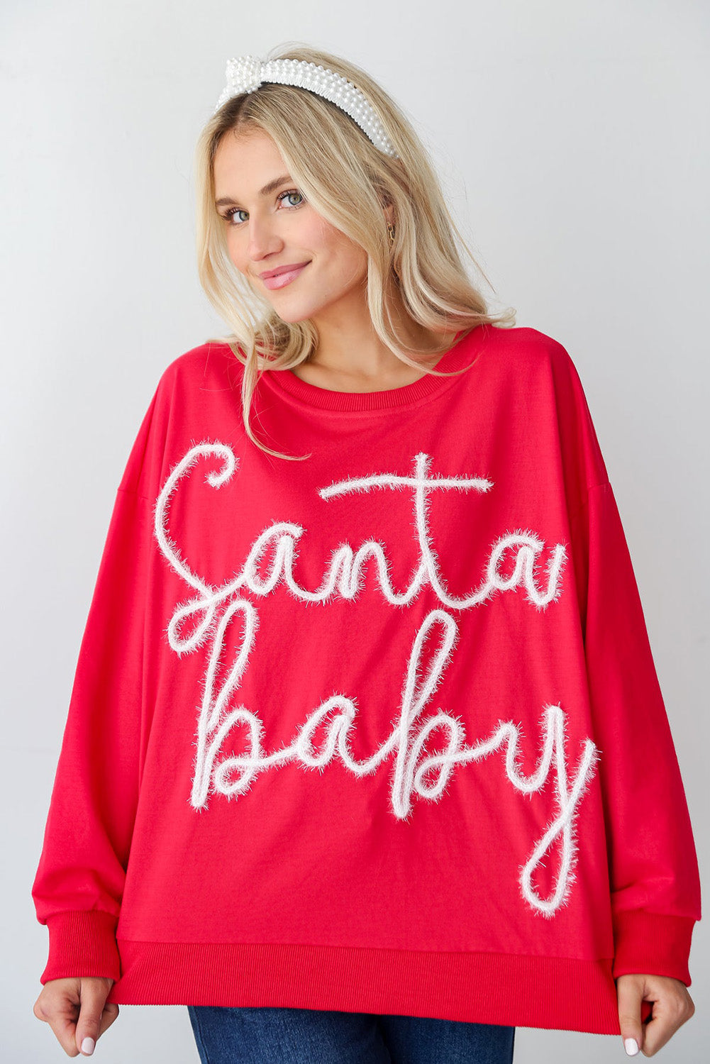 A person with long blonde hair is seen wearing the Fiery Red Christmas Santa Baby Tinsel Graphic Sweatshirt and blue jeans, facing away from the camera against a plain background.
