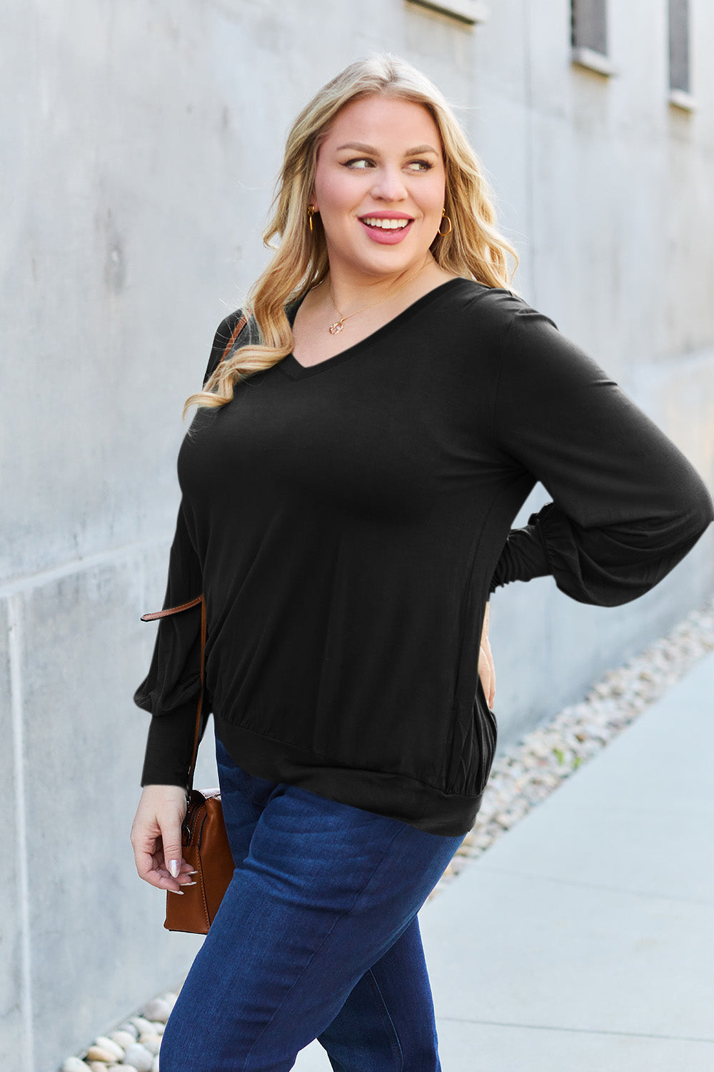 A woman with brown hair is standing against a concrete wall, wearing the Basic Bae Full Size V-Neck Lantern Sleeve Top in teal and blue basic-style jeans, smiling.