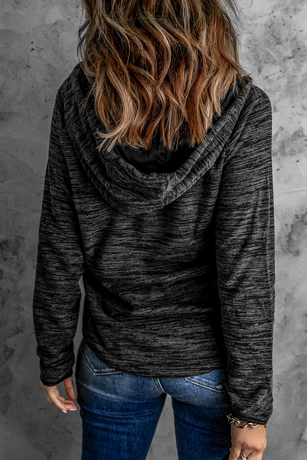 Person with wavy hair, wearing a stylish Black Heathered Print Button Snap Neck Pullover Hoodie and blue jeans, standing with their back facing the camera against a gray textured wall.