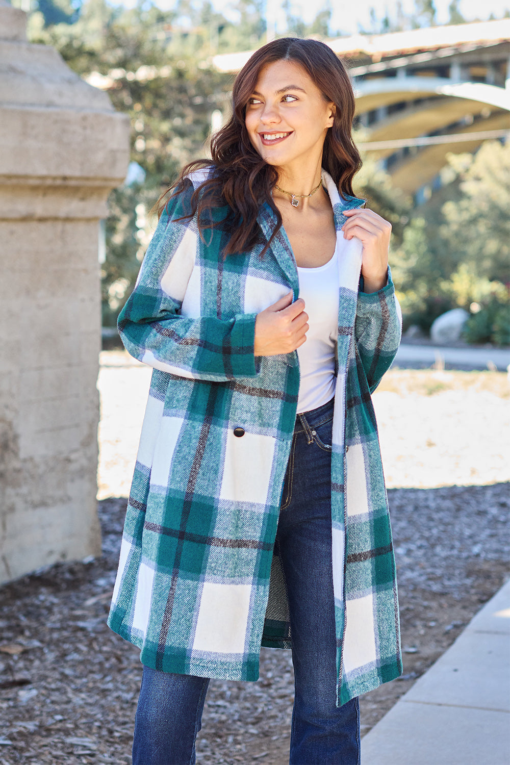 A woman stands on a sidewalk wearing the Double Take Full Size Plaid Button Up Lapel Collar Coat in pink and white over a white top and blue jeans, with her right hand lightly touching her hair. The polyester fabric of the coat makes it durable and perfect for an easy machine wash cold.