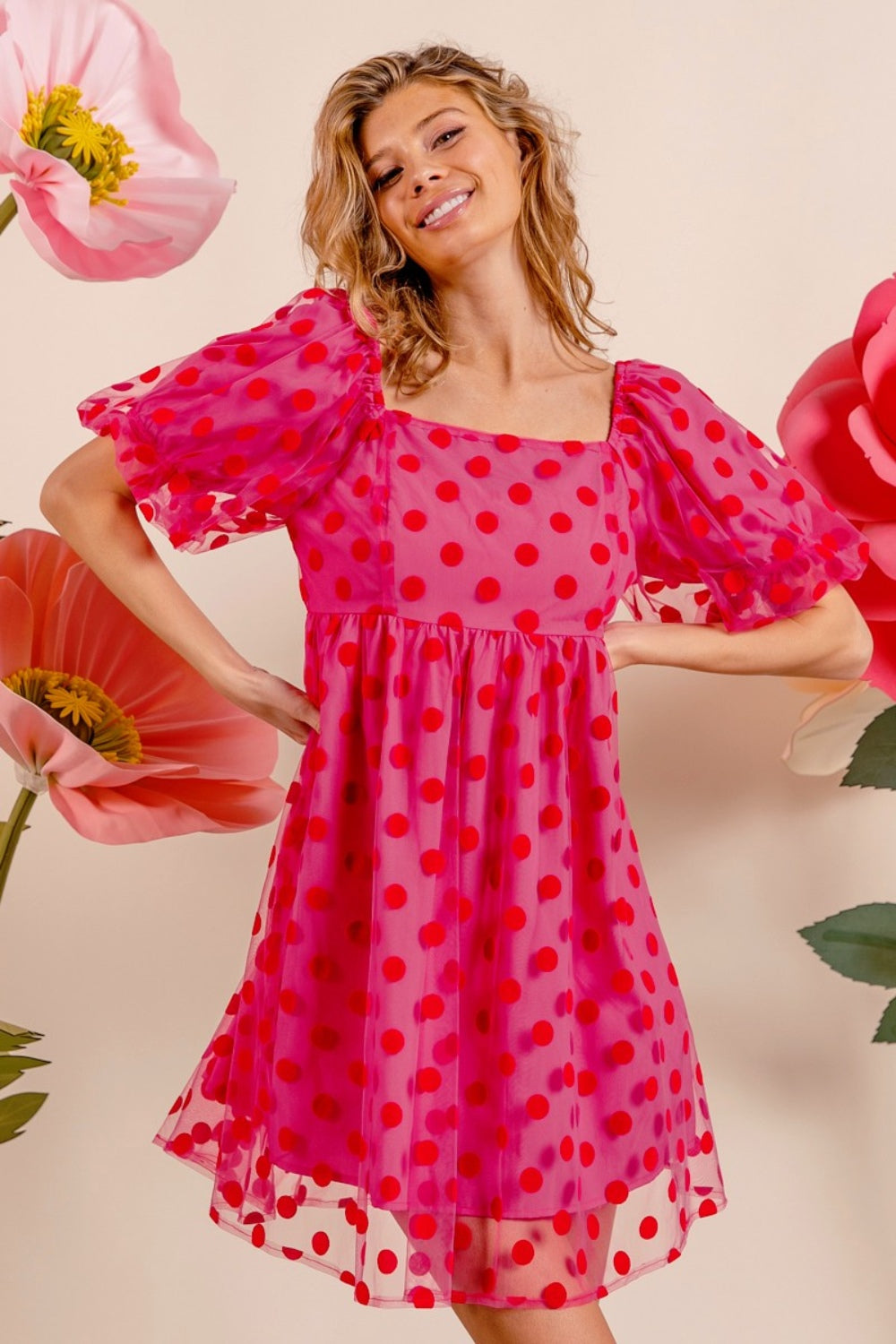 Wearing the BiBi Polka Dot Mesh Puff Sleeve Dress, a woman poses gracefully with one hand on her head. The vintage-inspired pink dress adorned with red polka dots enhances her elegance as she stands amidst large artificial flowers.